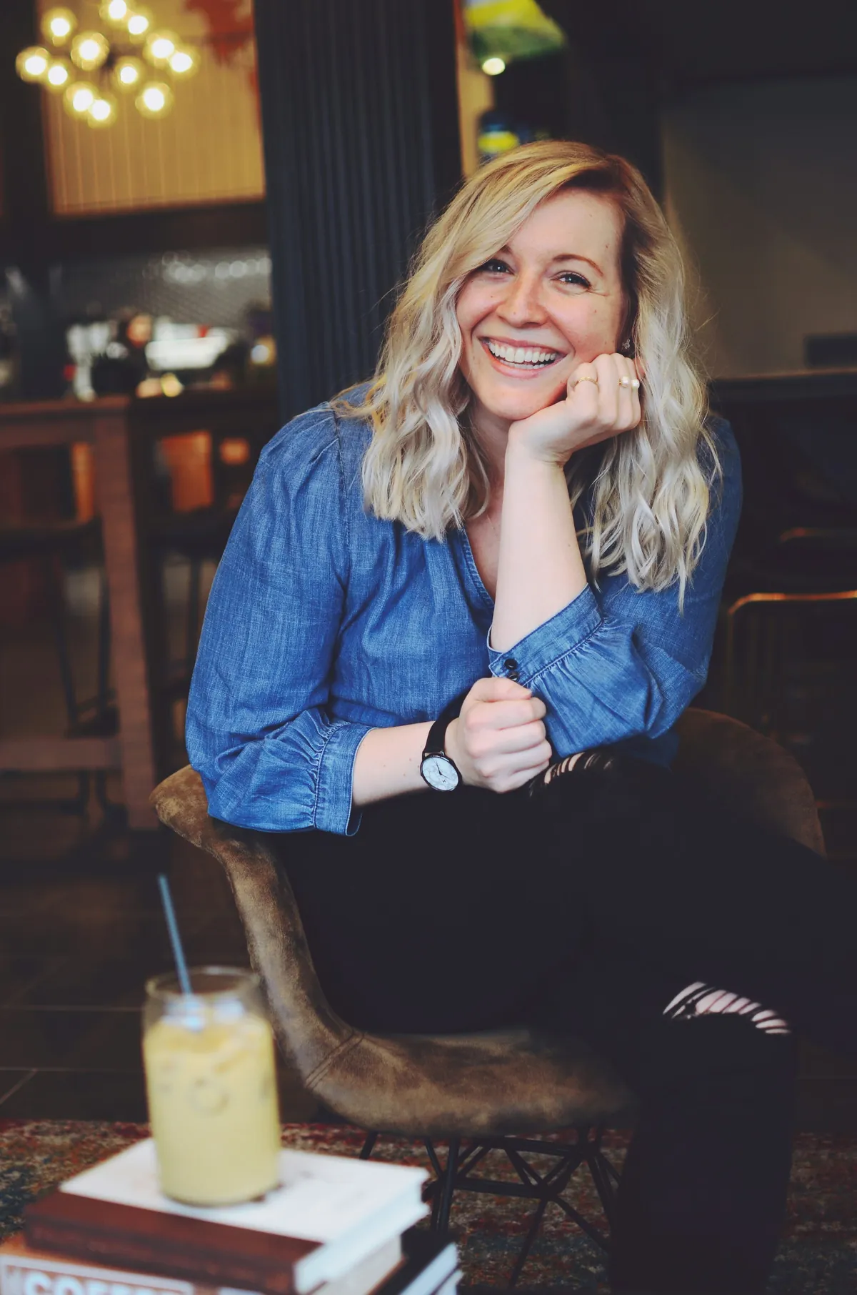A smiling woman with blonde curly hair wearing a blue denim shirt, sitting on a chair in a co-working space, with a yellow drink with a straw on the table in front of her.