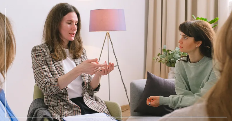 Group therapy session with five women, two visible, engaged in a discussion in a cozy room with a lamp and plants.