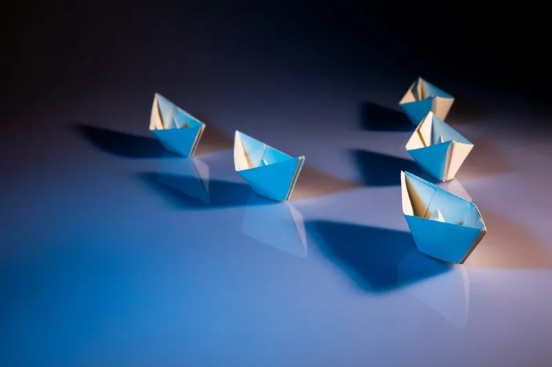 Six small paper boats arranged on a reflective surface with blue and purple lighting, casting shadows, representing leadership.