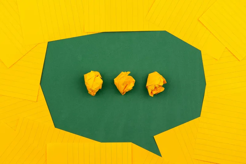 Three crumpled yellow paper balls on a green speech bubble-shaped paper, surrounded by additional yellow lined paper sheets.