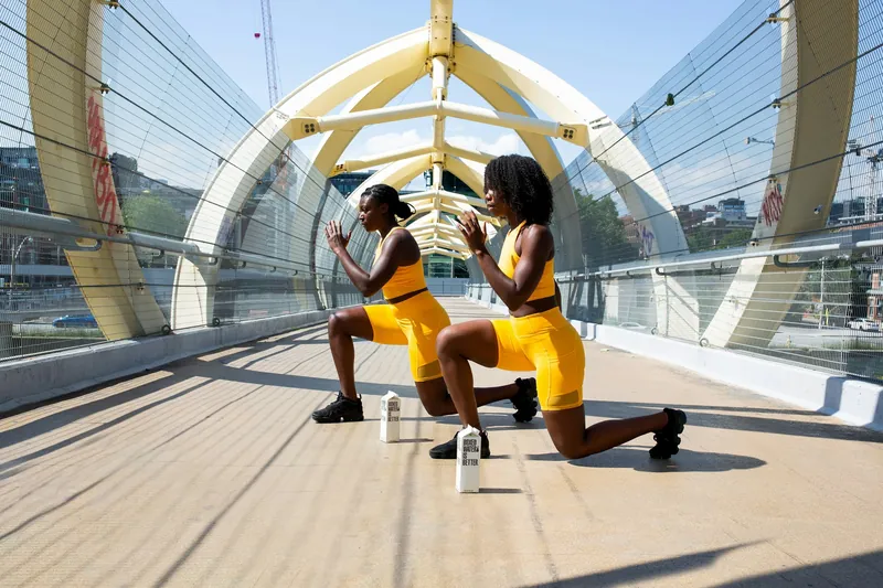 Two women practicing yoga on a bridge, kneeling on one knee with hands pressed together in prayer position, wearing yellow athletic wear, with a water bottle nearby and city buildings in the background.